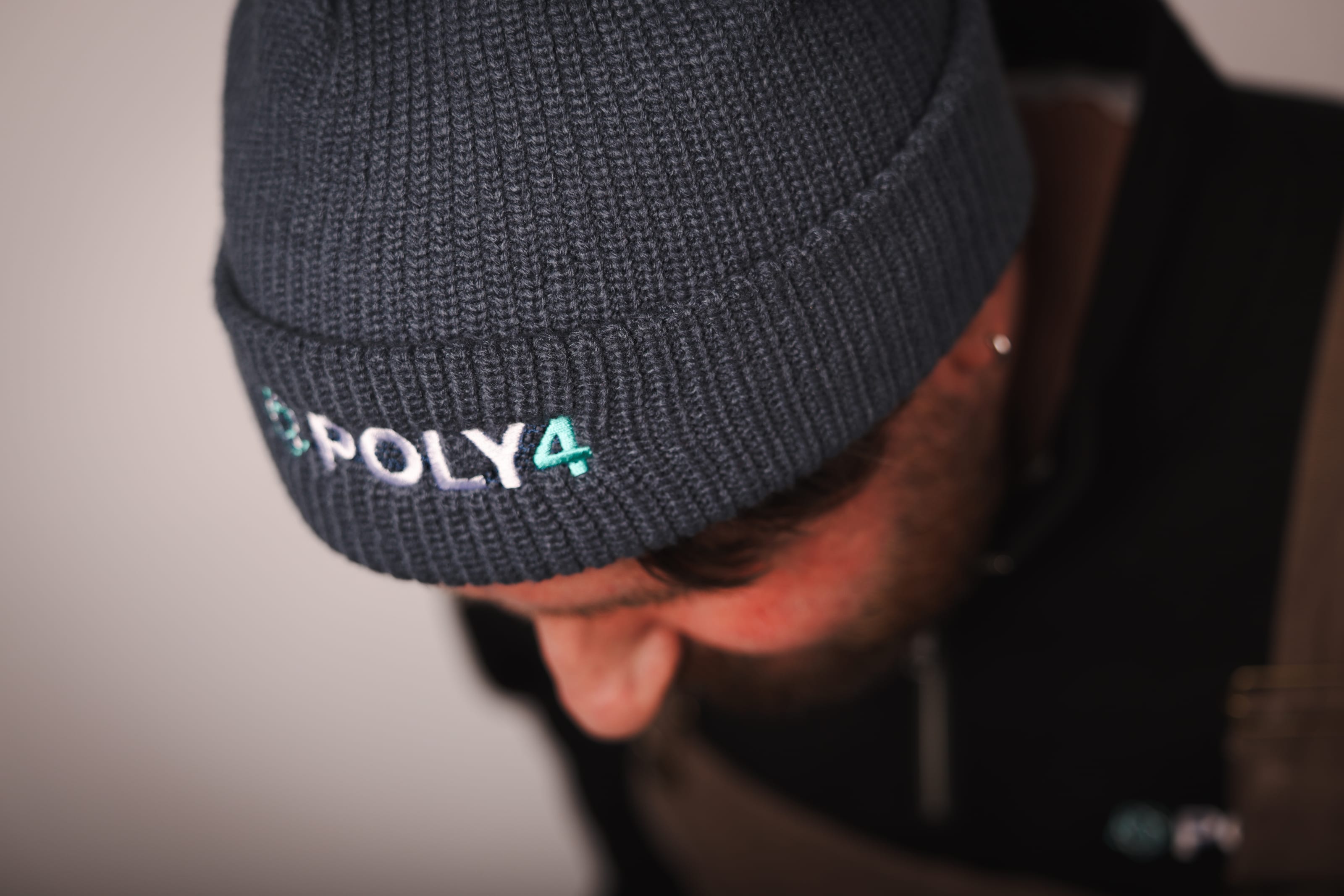 Close-up of POLY4 embroidered in white and teal on a charcoal ribbed beanie.